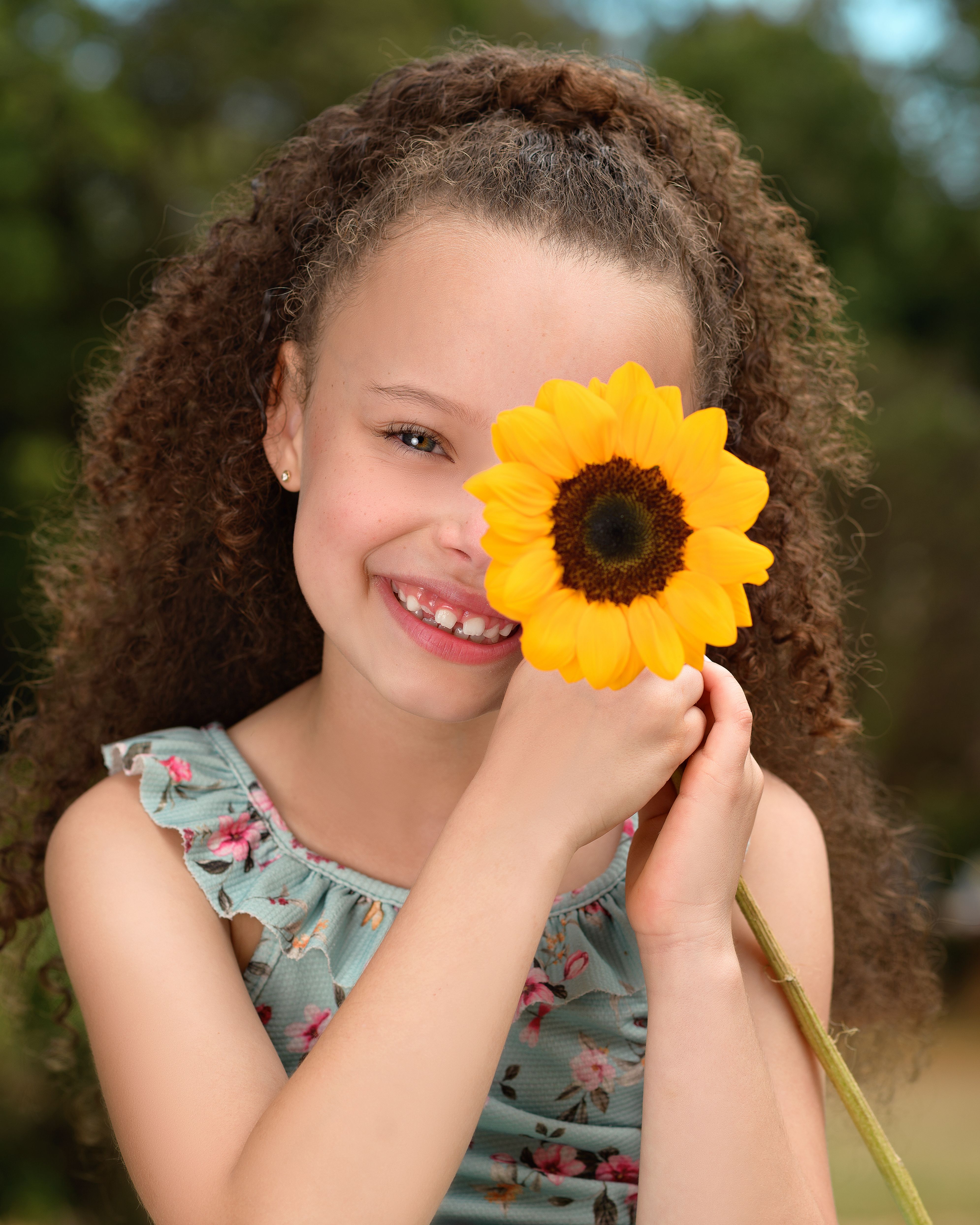 Smiling girl with curly hair holding a vibrant sunflower outdoors in summer.