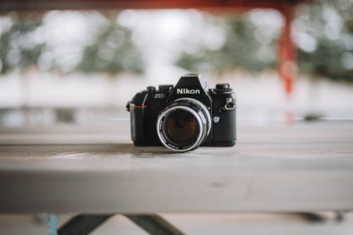 Free Close-up of a vintage Nikon film camera resting on a wooden table outdoors, surrounded by soft bokeh. Stock Photo