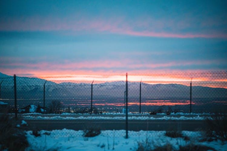 Fences Separating Road From Snowy Fields At Sunset