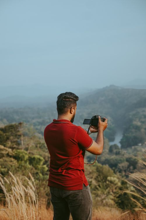Free A man capturing a scenic mountain view with his camera, surrounded by lush greenery. Stock Photo