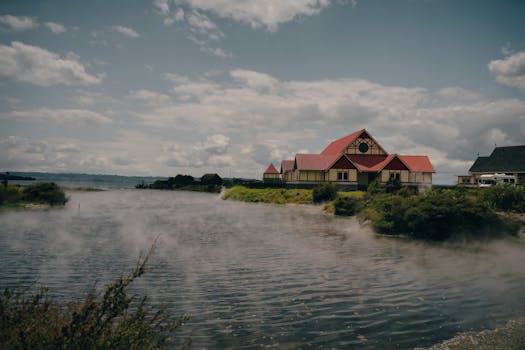 Traditional Maori building by Rotorua Lake with geothermal steam rising, captured in daylight.