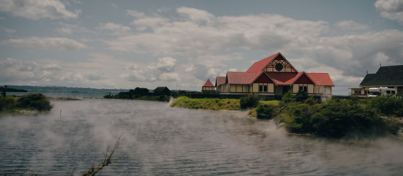 Traditional Maori building by Rotorua Lake with geothermal steam rising, captured in daylight.