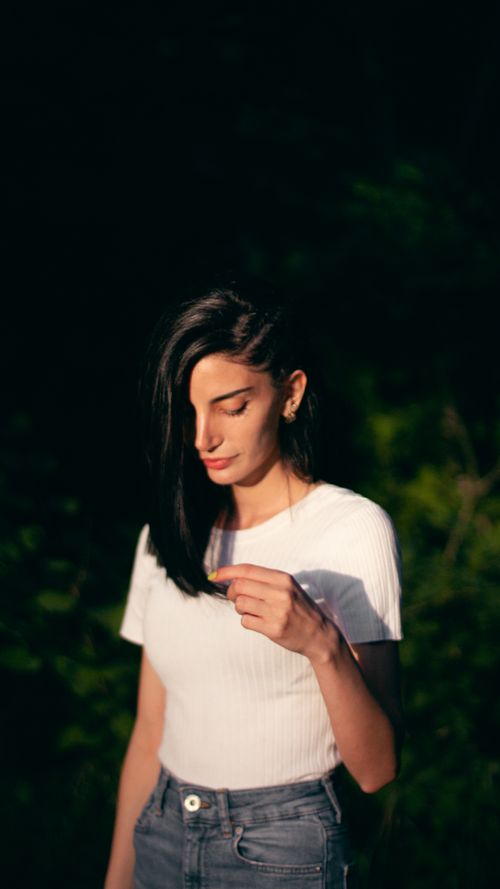 Free A woman with black hair stands outdoors wearing a white top and denim jeans, bathed in sunlight. Stock Photo