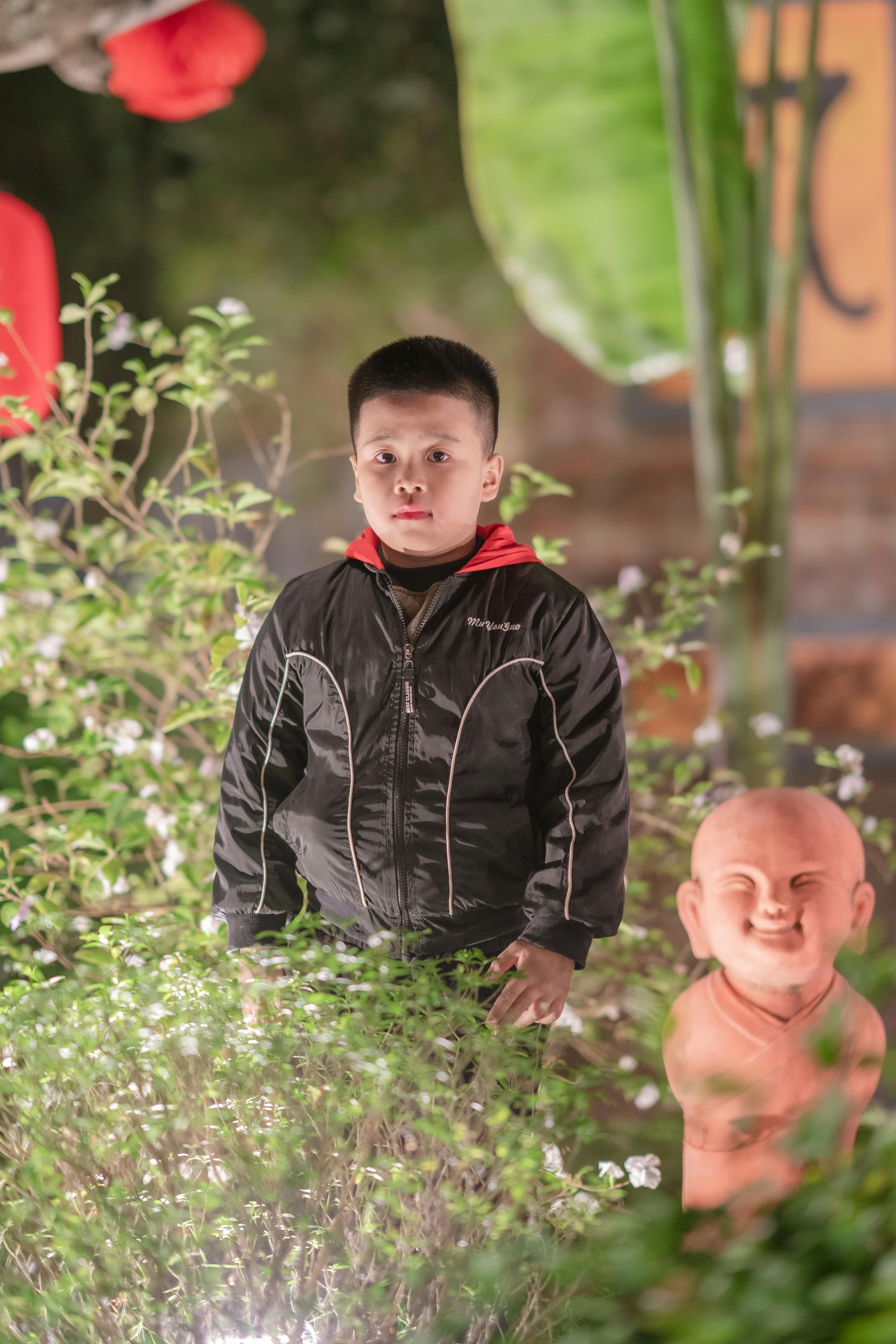 A boy stands amidst lush greenery and a smiling statue in a garden setting.