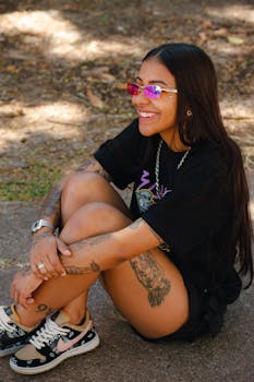 Teenager sitting outdoors, showcasing tattoos and wearing stylish sunglasses with a bright smile.