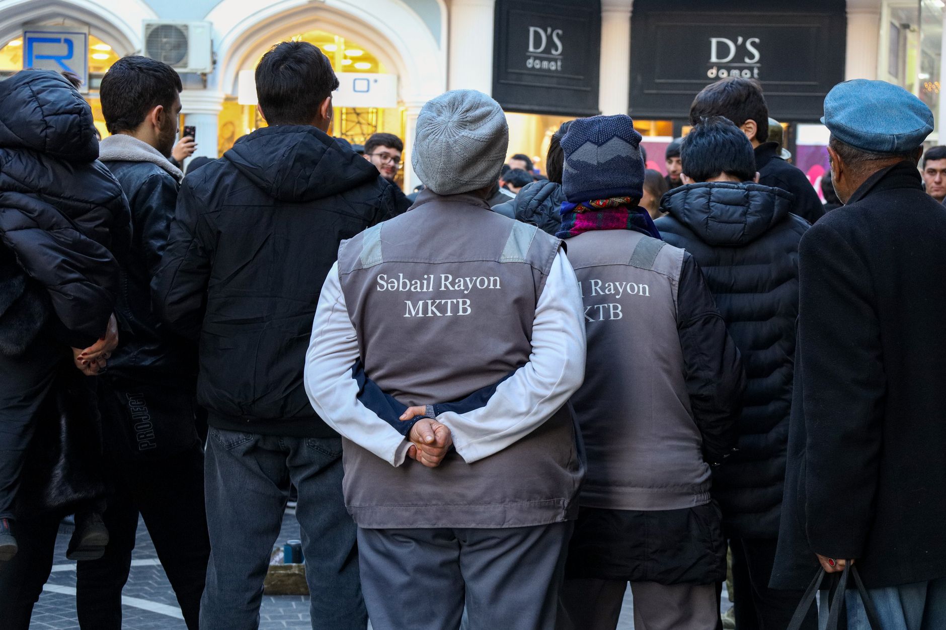 A diverse crowd standing on a busy street, featuring people in uniforms and knit hats during the daytime.