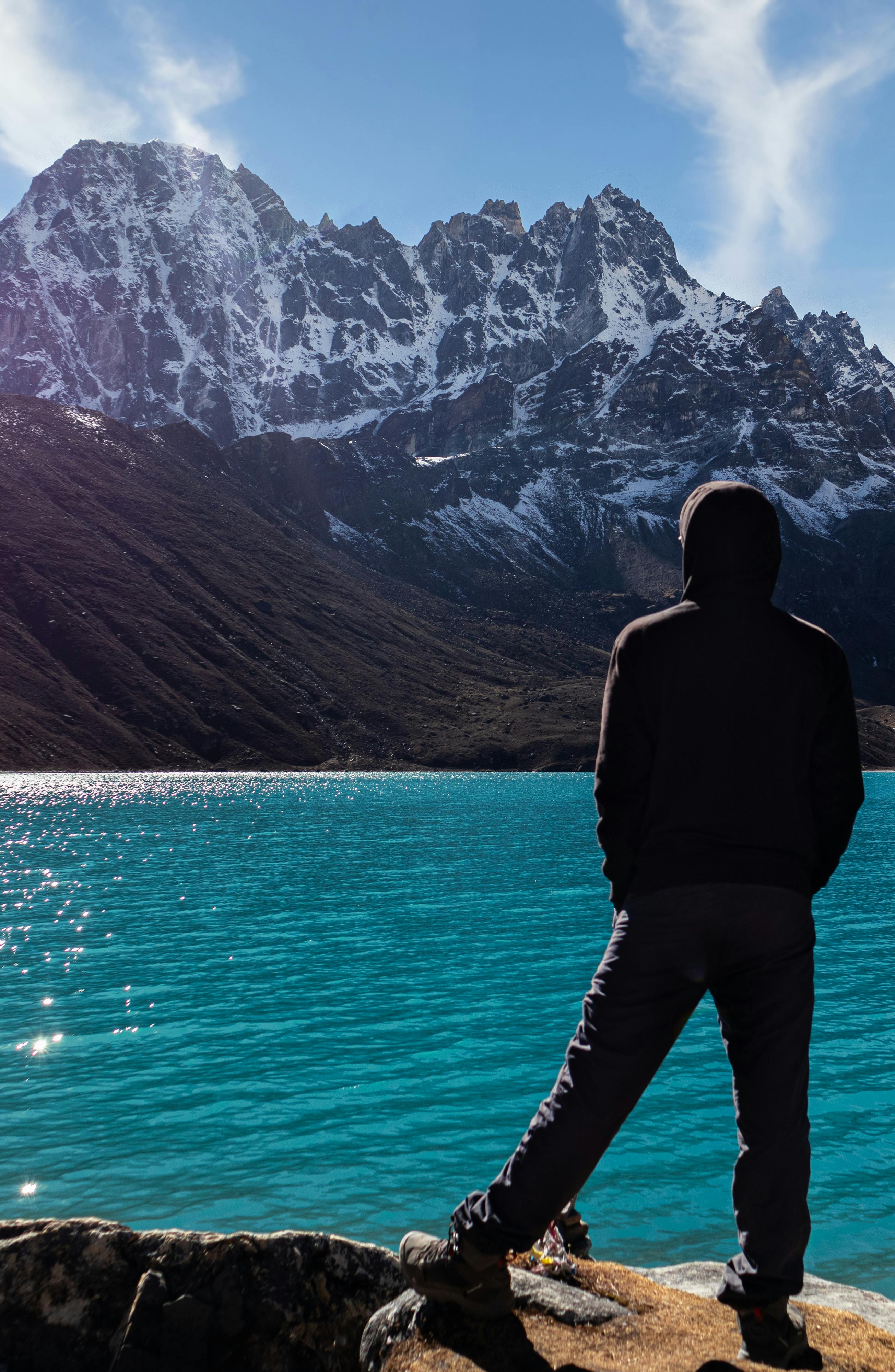 a silhouette of a man in nepal is overlooking the gokyo village s turquoise blue lake into the blue and cloudy sky with a big snow capped mountain in between