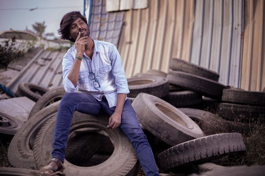 A thoughtful man with dark hair poses outdoors on stacked tires wearing a light shirt and blue pants.