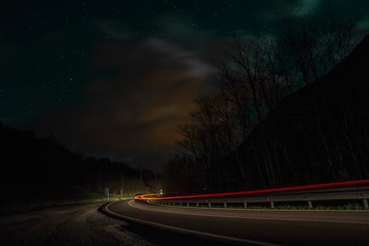 Dynamic car light trails on a curving road at night with a starry sky and clouds.