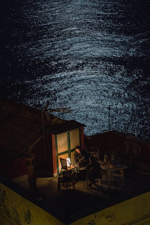 Men Sitting on House Roof on Lakeshore at Night