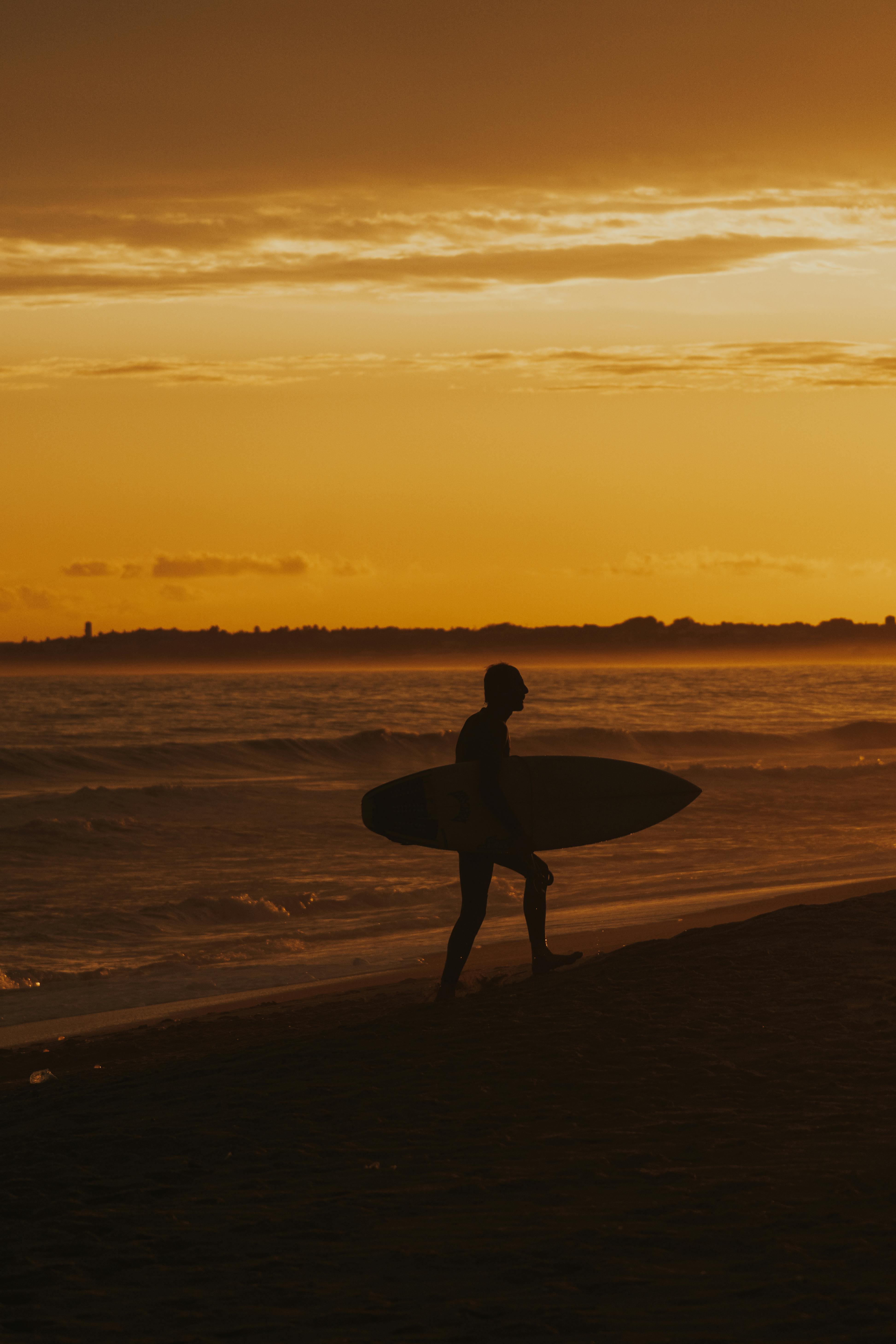 silhouette of a man carrying his surfboard while walking on the beach during sunset