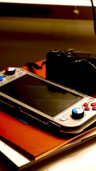 A portable gaming console rests on a stack of notebooks with a camera in the background.