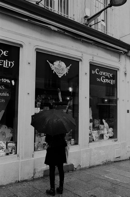 Black and White Photo of Person with Umbrella Looking at Display Window of Store