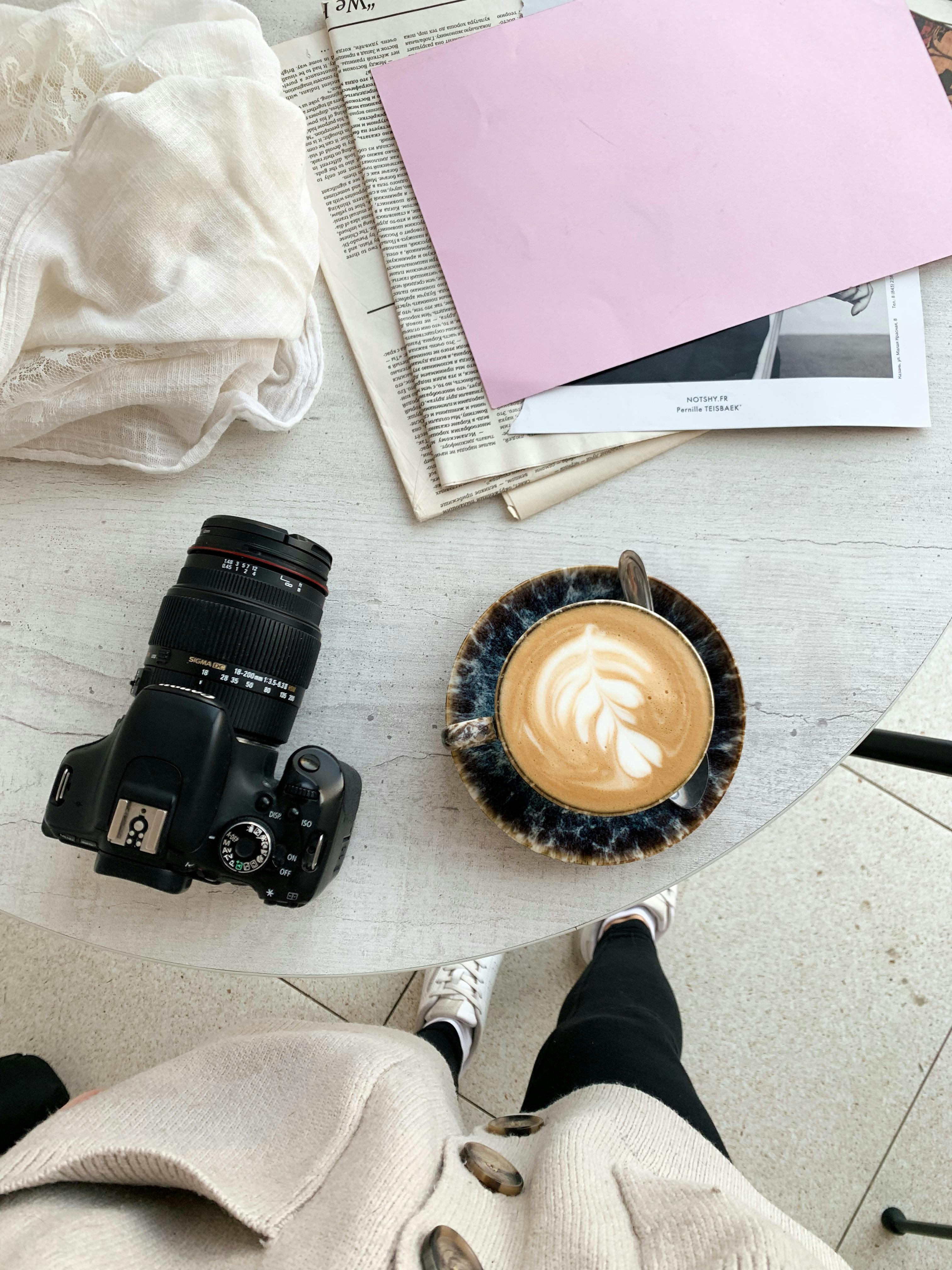 Overhead shot of a coffee cup with latte art, DSLR camera, and papers on a round table.