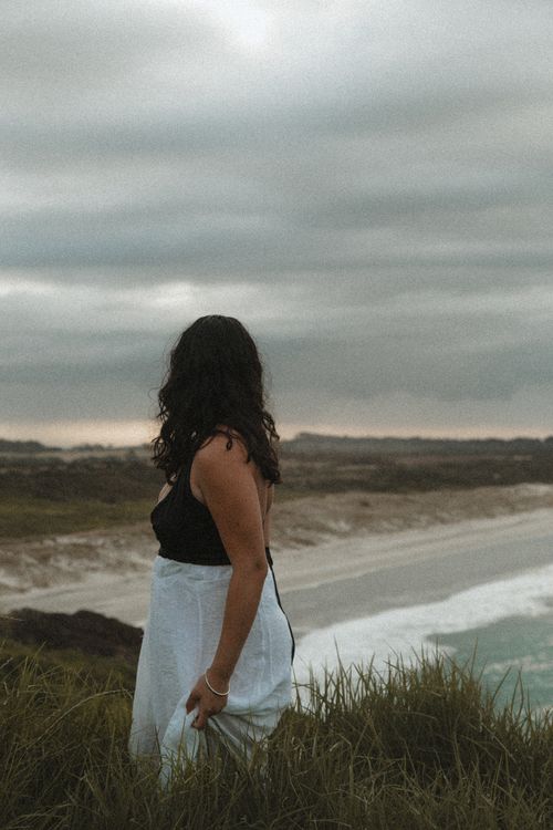 Free A woman gazes towards the sea on a cloudy beach day, creating a serene moment. Stock Photo