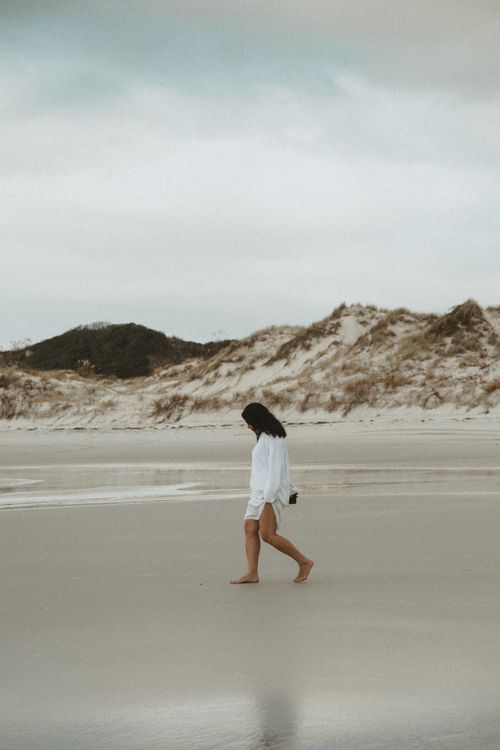 Free A woman strolls peacefully along a sandy beach with scenic dunes in the background. Stock Photo