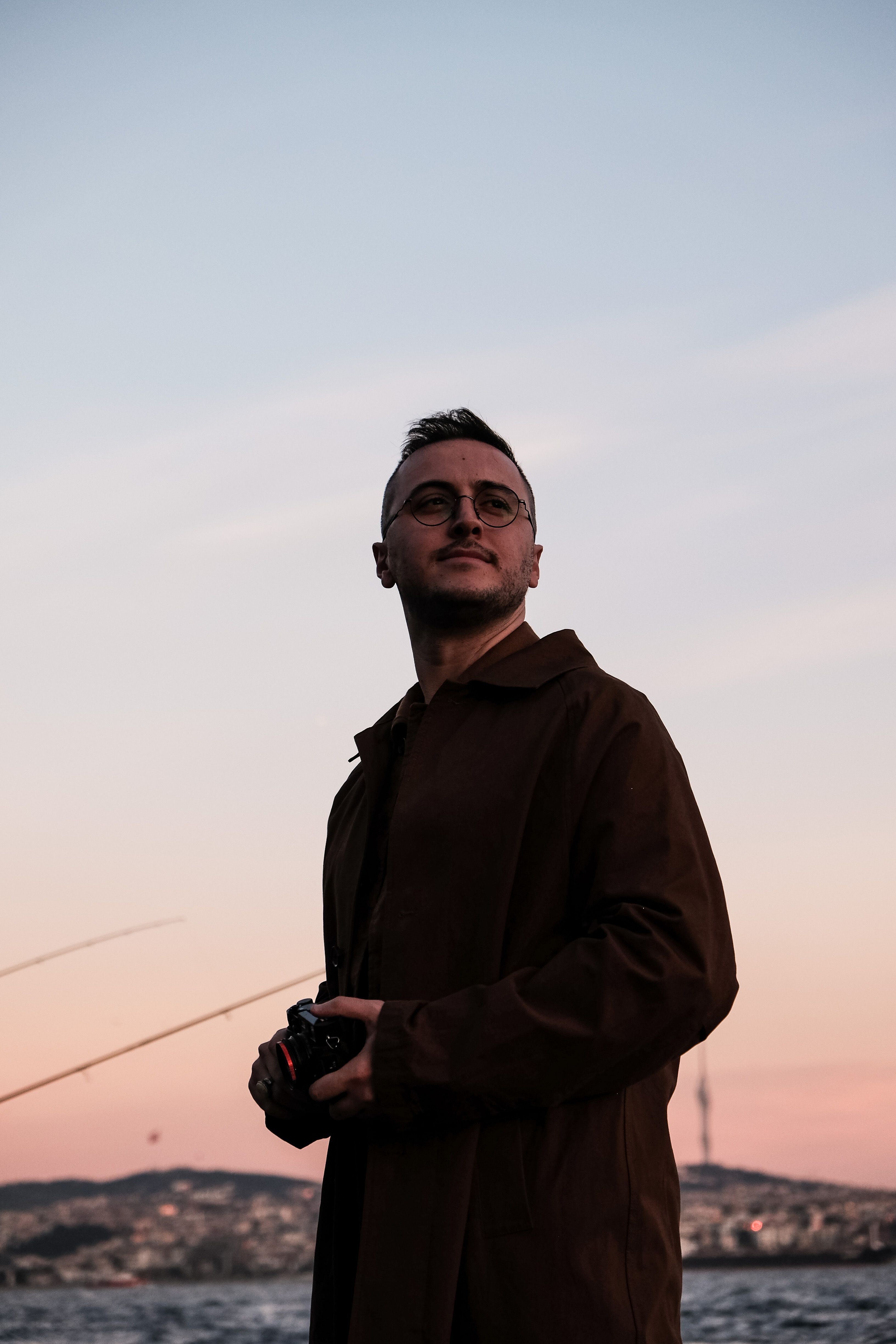 A man with eyeglasses stands with a camera by the sea during twilight, against a serene cityscape backdrop.
