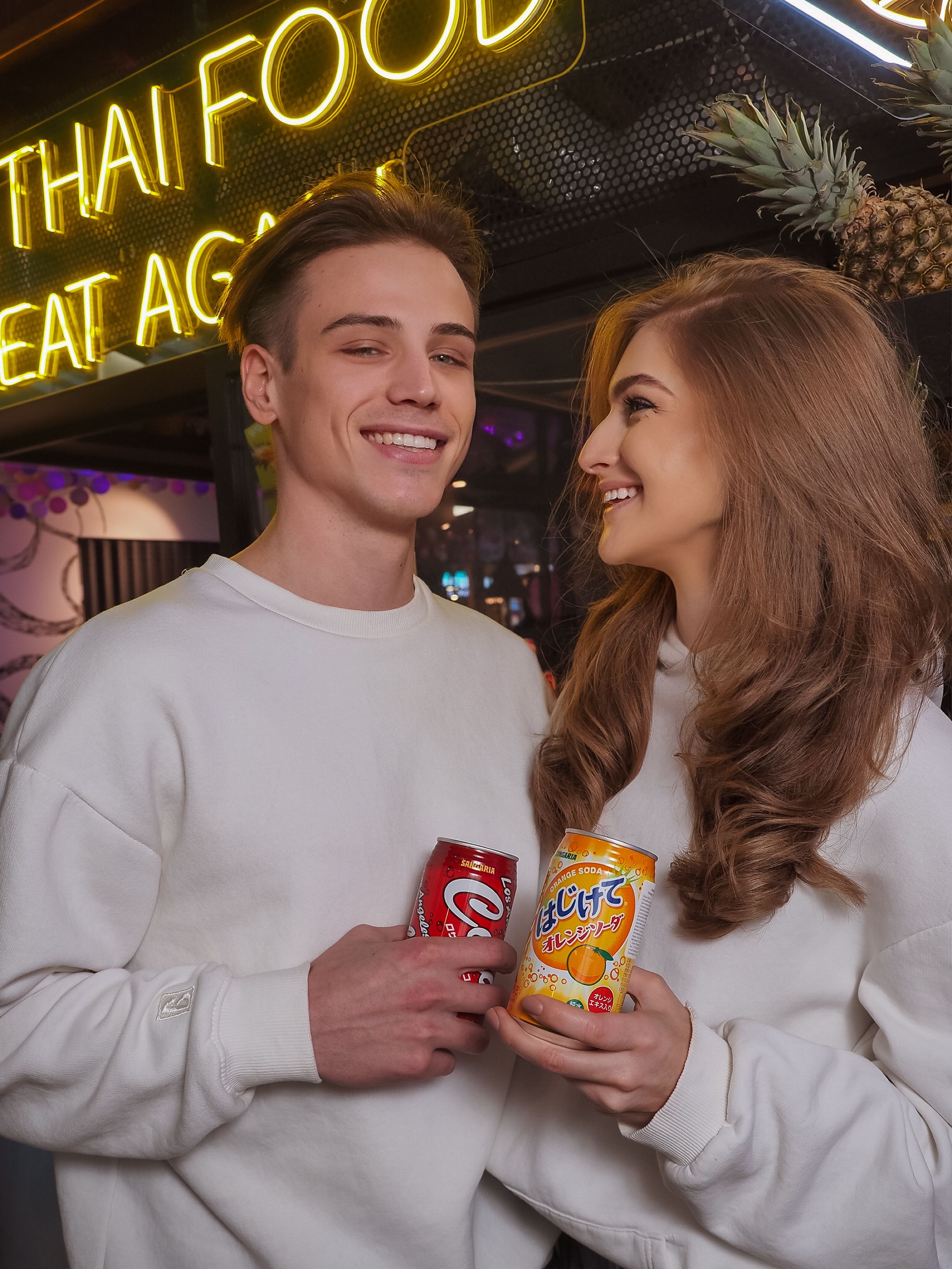 Smiling couple holding soda cans at a neon-lit restaurant, creating a lively atmosphere.