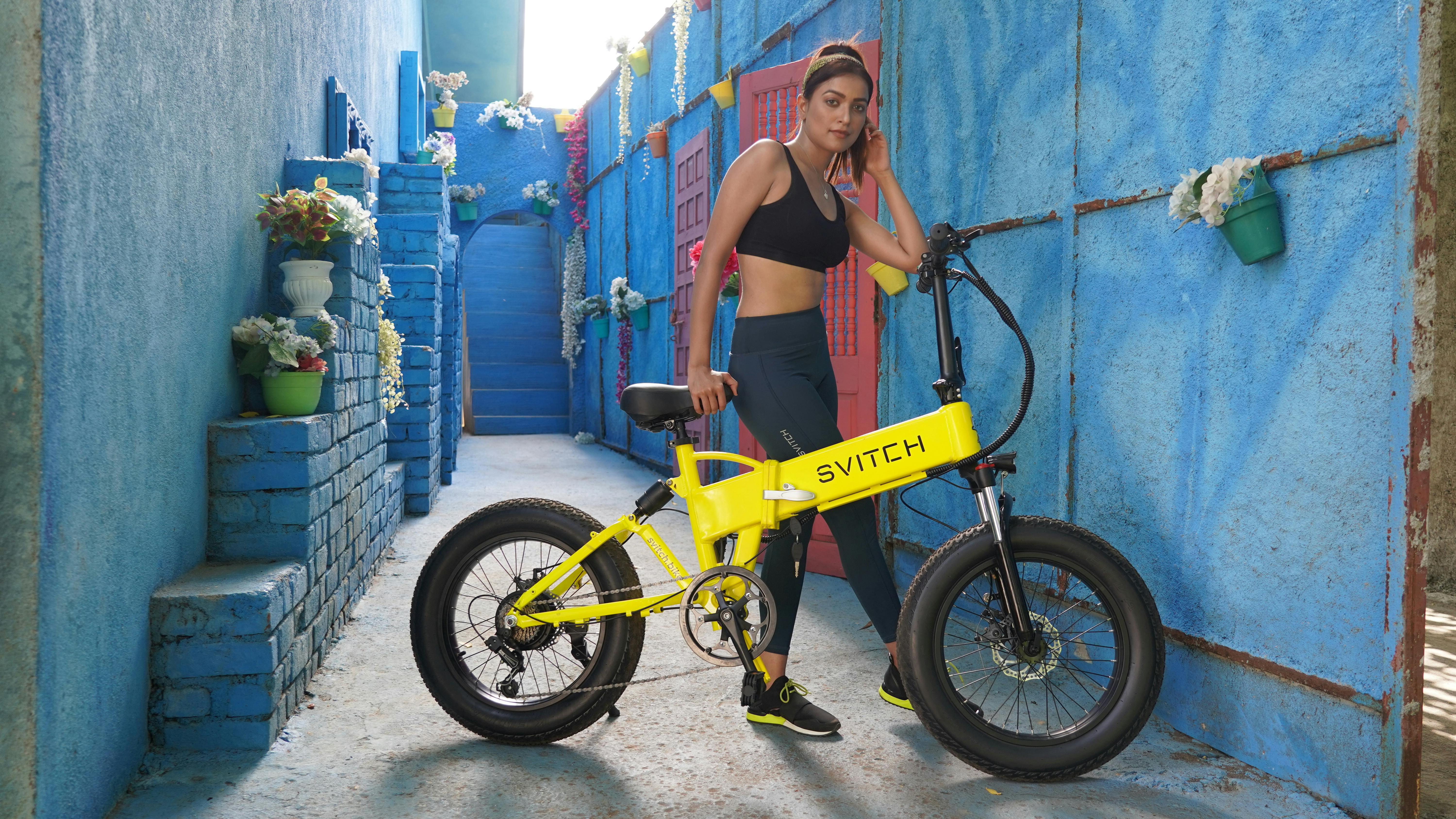 Free A woman in activewear standing with a yellow electric bike against a colorful outdoor backdrop in India. Stock Photo