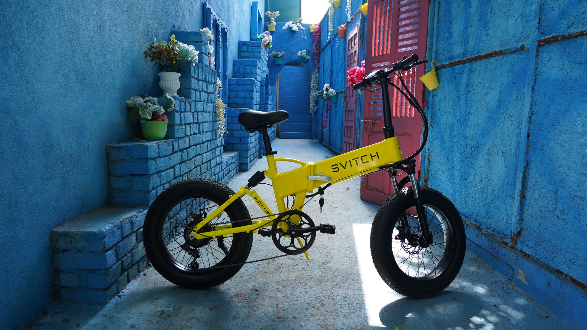 A stylish yellow foldable bicycle parked in a vibrant blue alley, adorned with flowers.