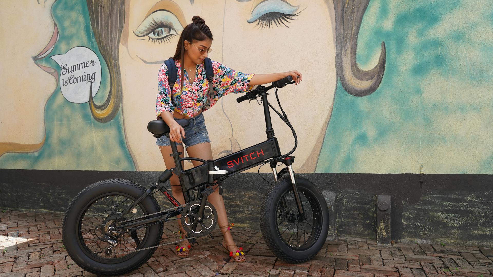 Stylish woman stands with e-bike against colorful mural in Surat, India.