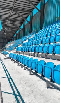 Brightly lit empty stadium with blue seats and metal structure, casting shadows.