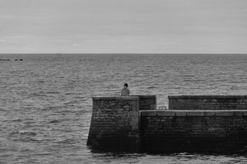 Free A lone person stands on a weathered stone pier gazing at the vast sea, captured in black and white. Stock Photo