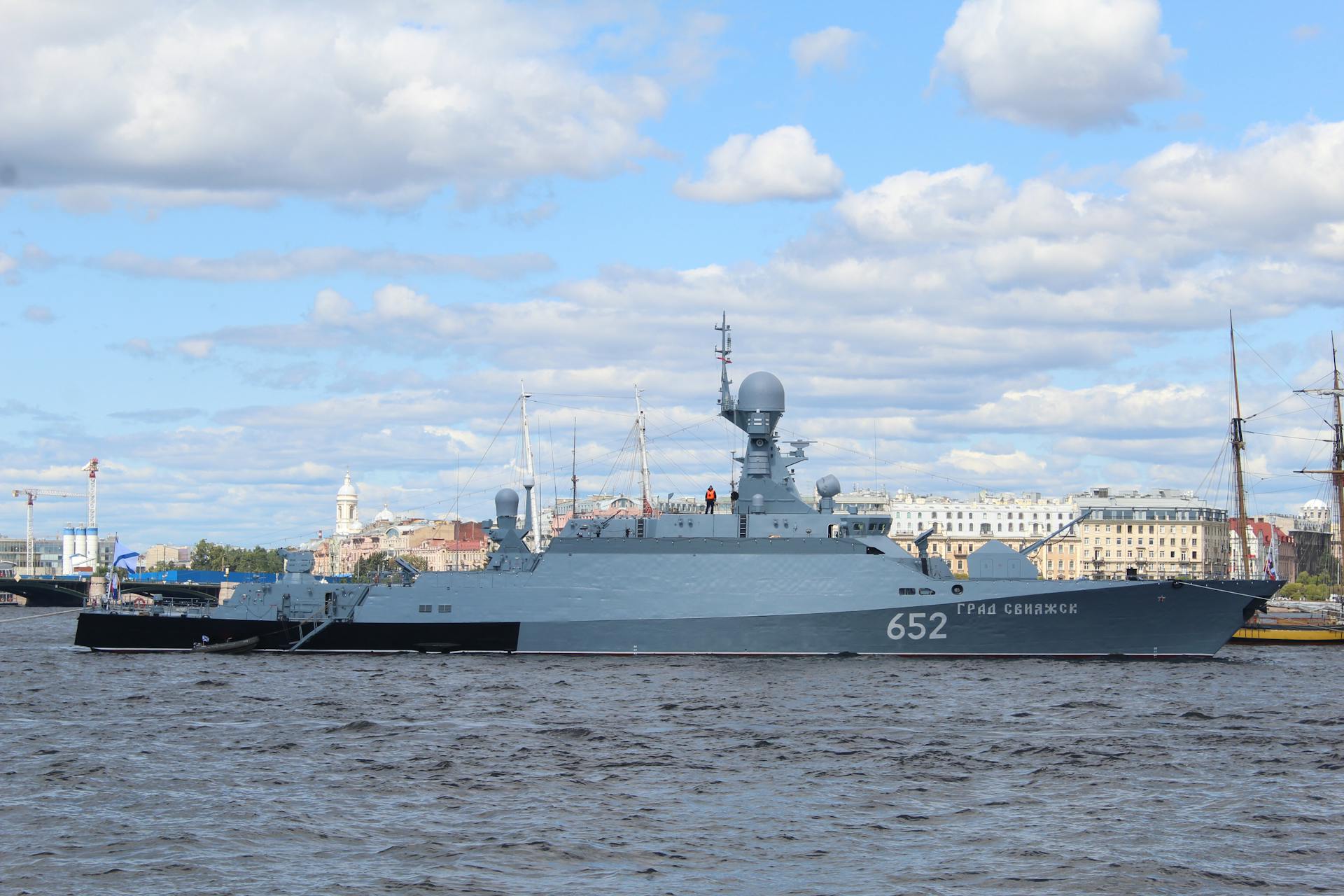 A modern navy warship docked in a European harbor on a clear day with cityscape background.