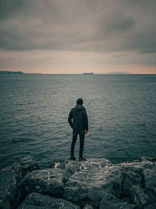 Free A man stands on rocks, gazing at the sea during sunset, evoking solitude and contemplation. Stock Photo