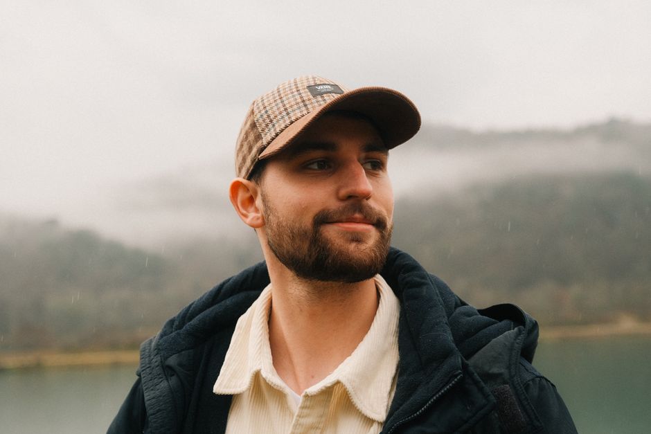 A bearded man in a cap outdoors, looking thoughtful in rainy weather.