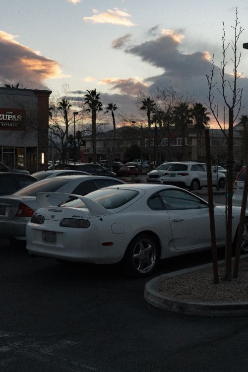 Free A white sports car parked in a lot during sunset with palm trees and vibrant clouds. Stock Photo