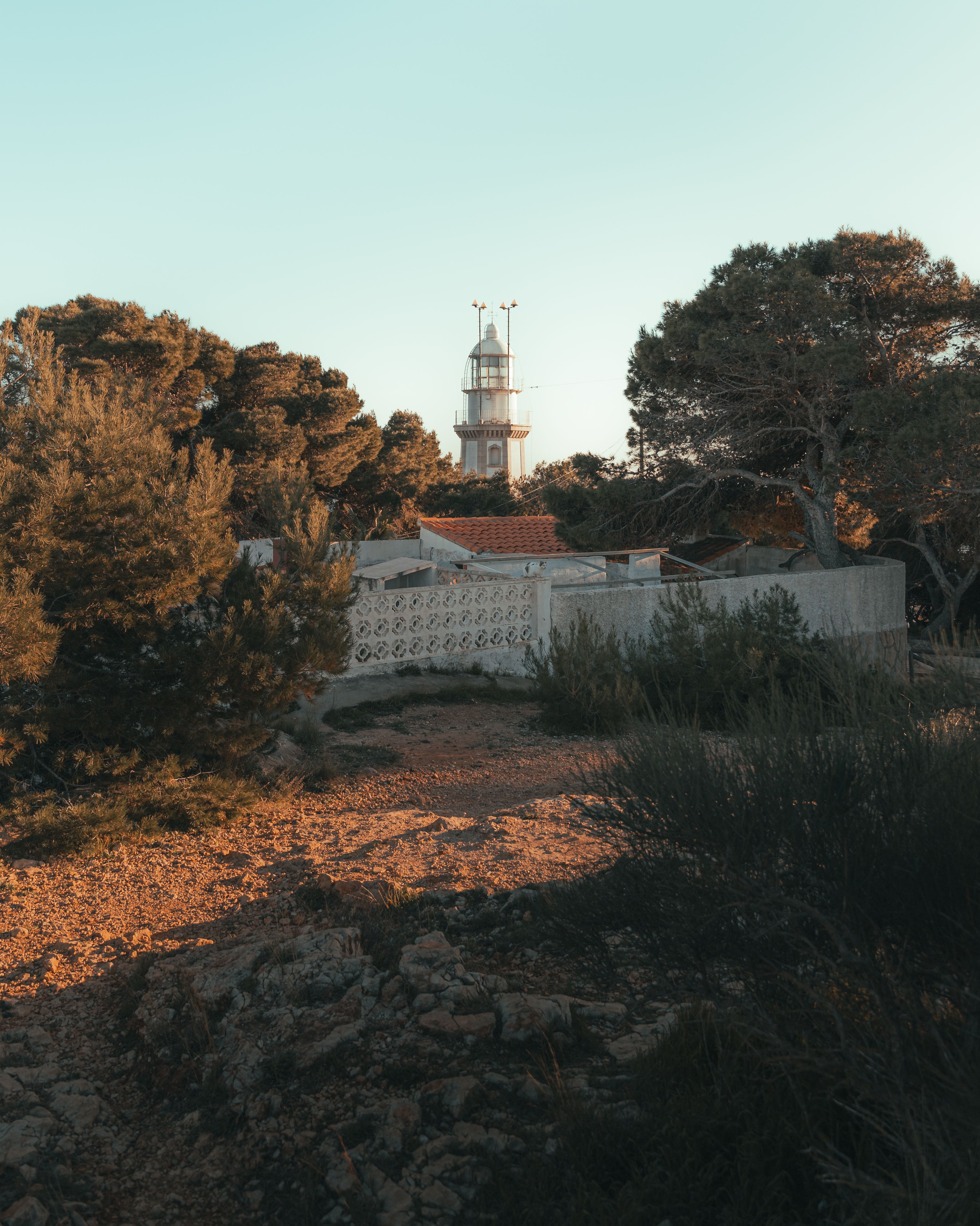 A serene coastal lighthouse surrounded by trees and a rocky path under a clear sky.