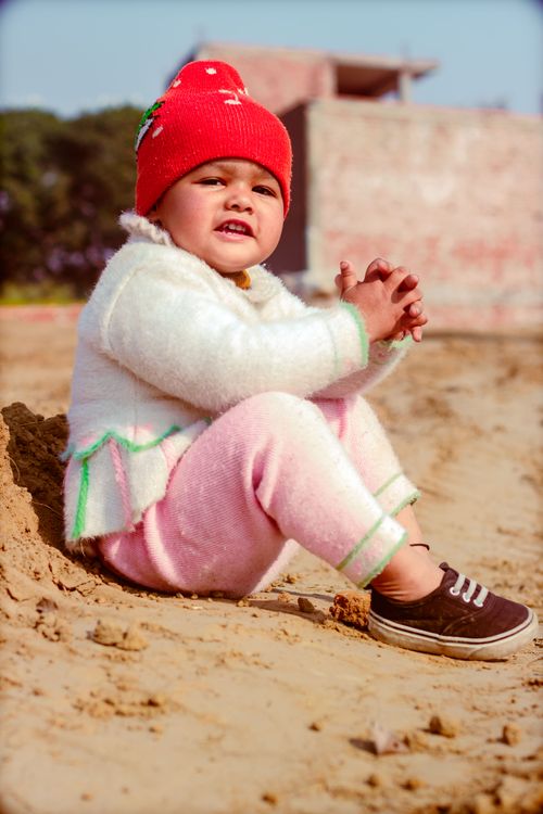 Free Charming toddler in winter clothing sitting on a sandy beach enjoying a sunny day. Stock Photo
