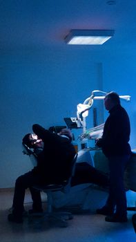 A dentist examines a patient in a dimly lit clinic room, silhouetted by blue light.