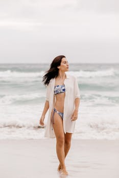 Woman in a bikini strolling along the sandy shore with waves in the background.