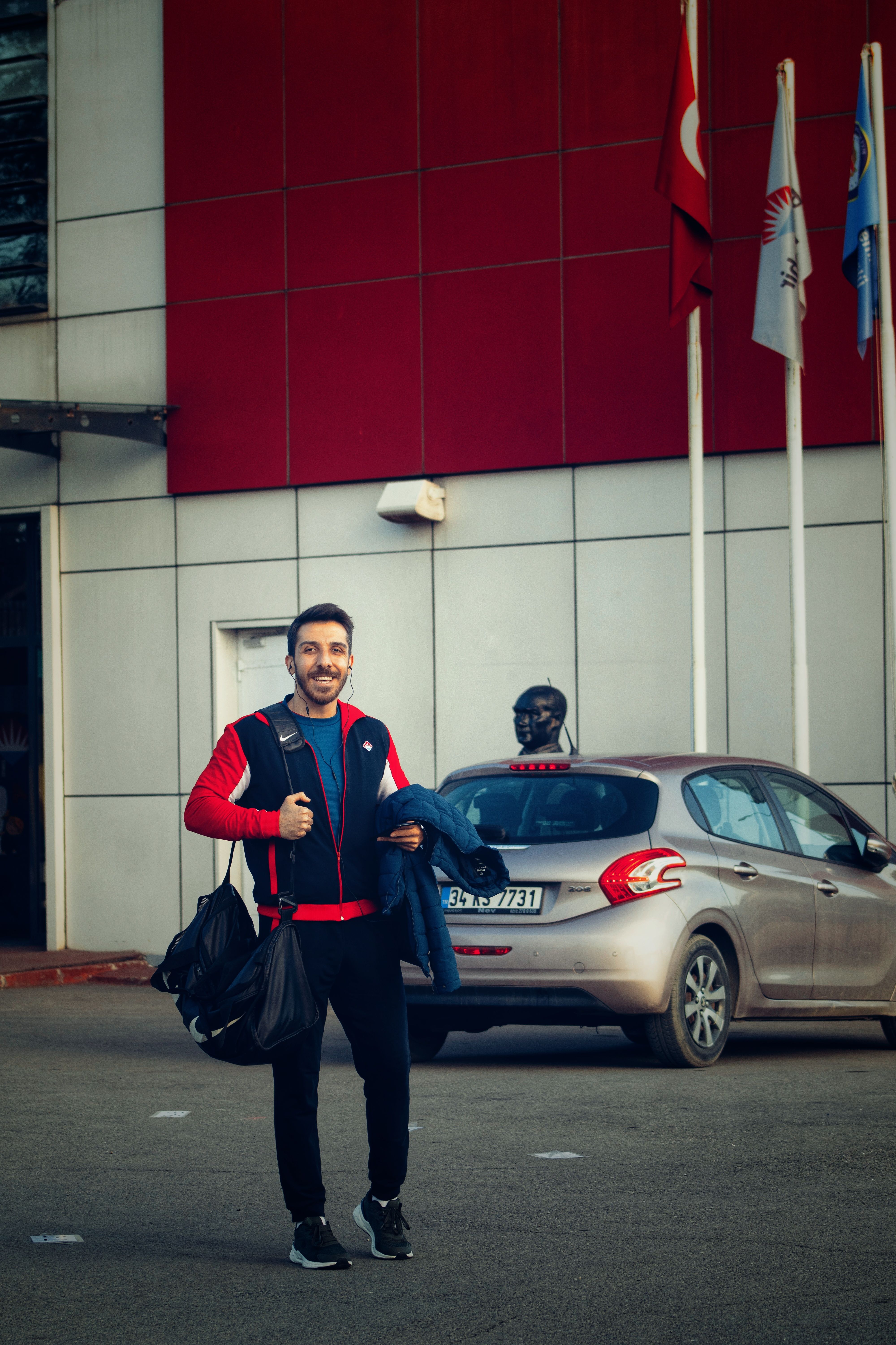 A man stands smiling outside a modern building, holding bags. Flags are visible, suggesting international or corporate environment.