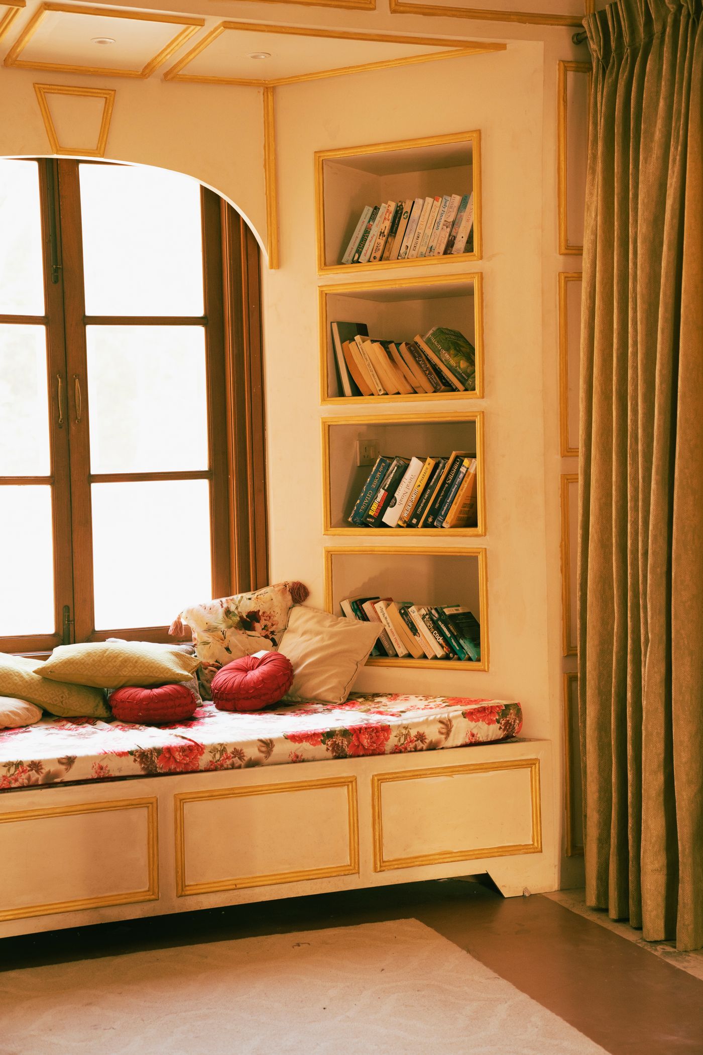 A built-in window-seat reading nook with floral cushions and floor-to-ceiling bookshelves beside a curtained window