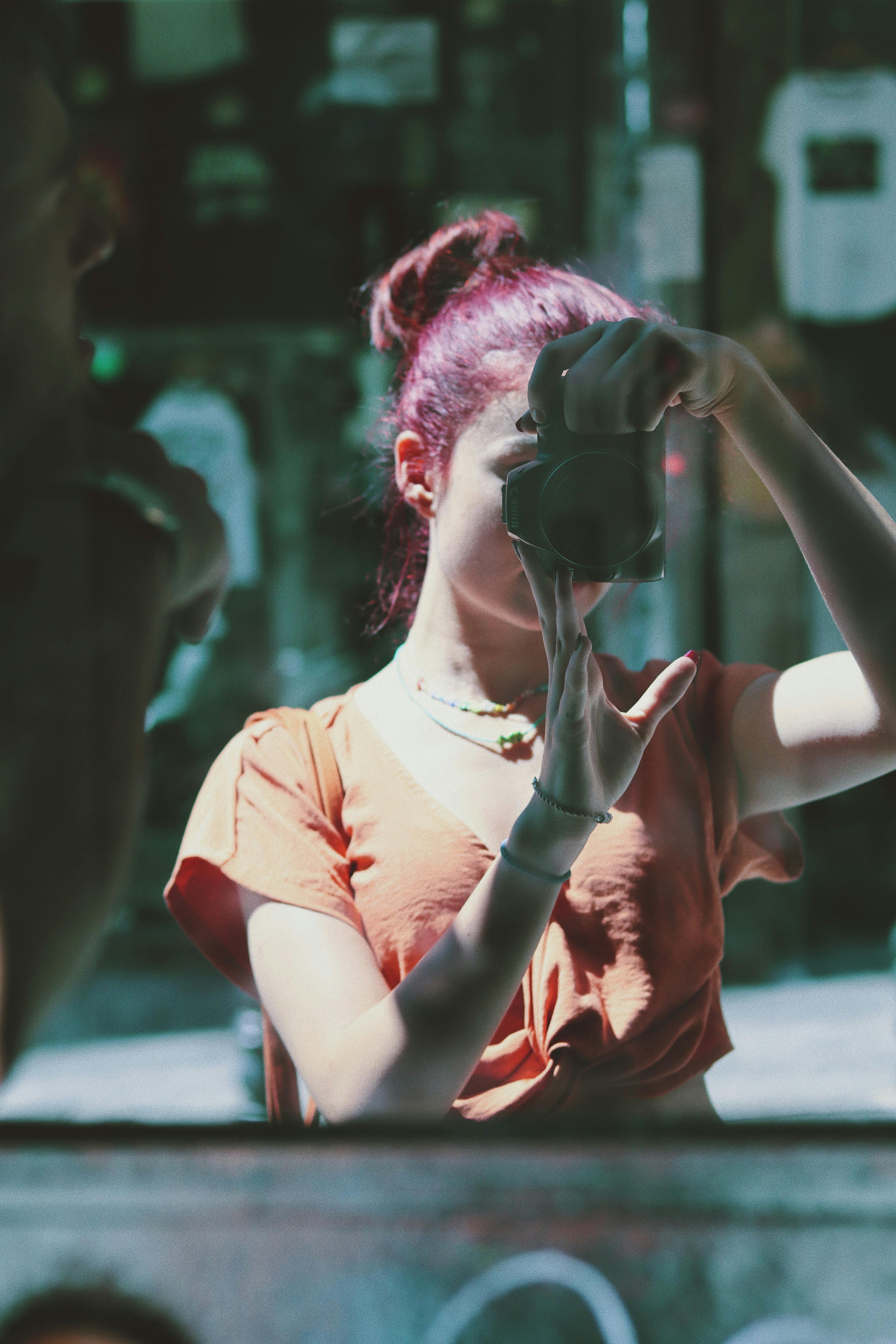 A young woman takes a mirror selfie outdoors, capturing her reflection with a camera.