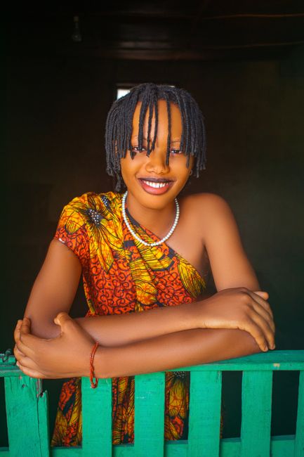 Vibrant portrait of a smiling woman in colorful traditional dress with braided hair leaning on a fence.