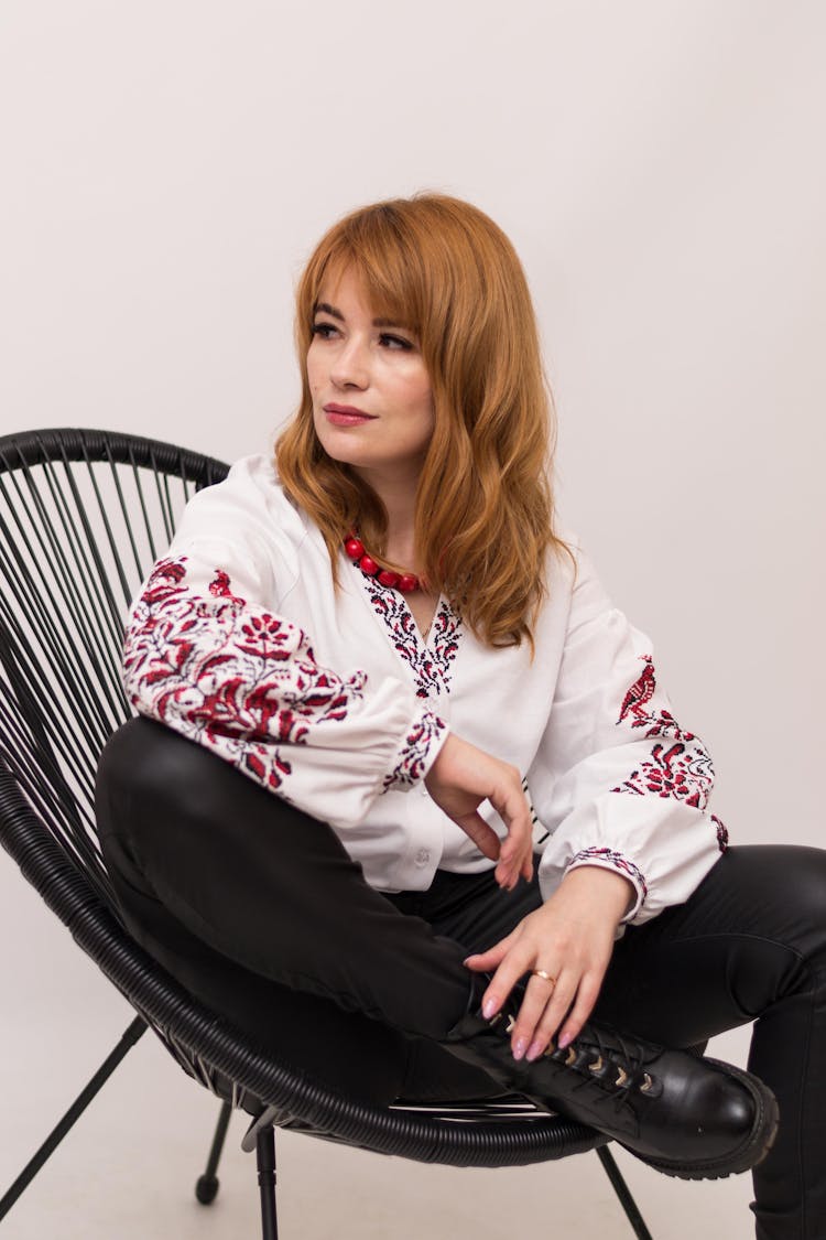 Redhead Woman In Embroidered Blouse Sitting In Chair In Studio