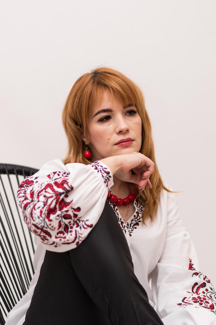 Redhead Woman In Embroidered Blouse Sitting On Chair In Studio