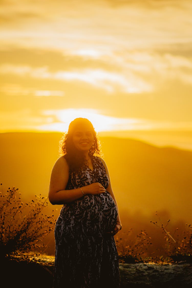 Pregnant Woman In A Dress Standing Outside At Sunset And Smiling 