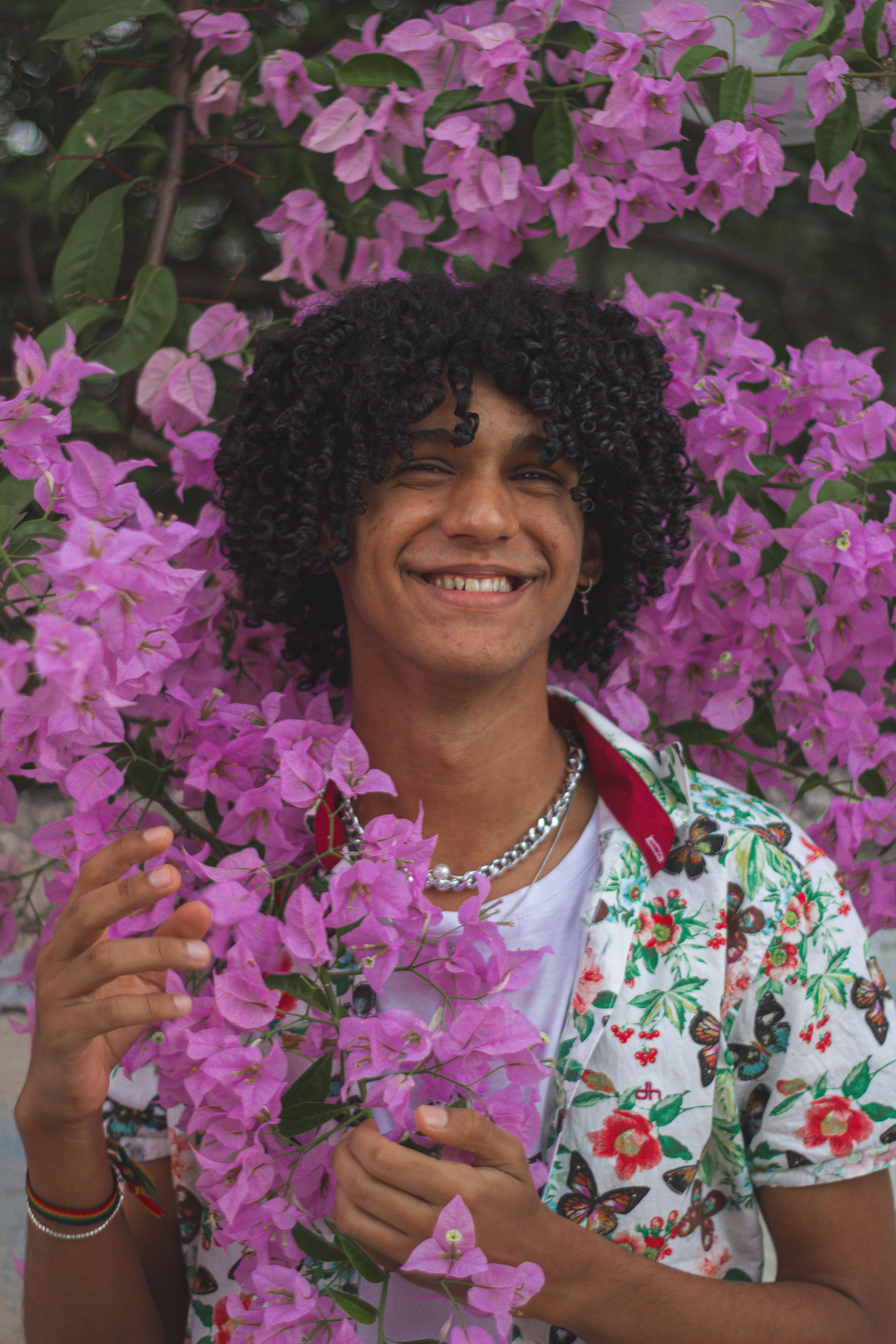 Cheerful man with curly hair poses among vibrant pink bougainvillaea blooms.