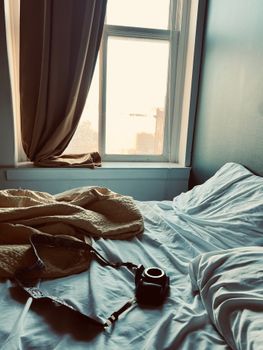 Sunlit bedroom with a camera on bed beside window, draped curtains.