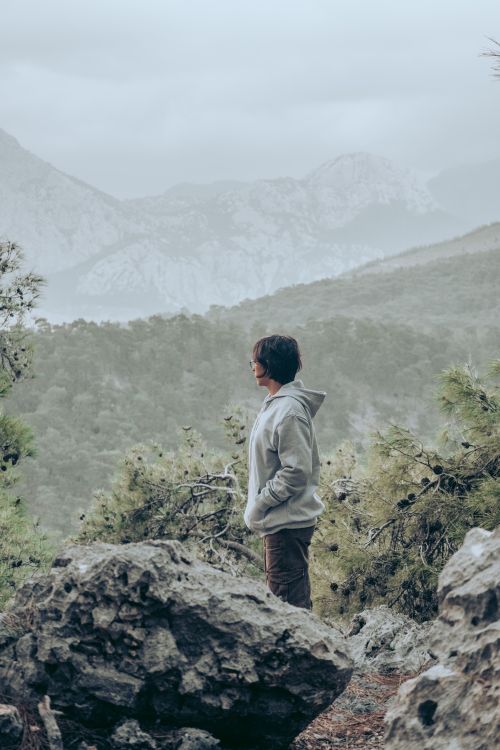 Free A woman in a gray hoodie stands amidst rocky terrain with mountains in the distance, embracing nature. Stock Photo