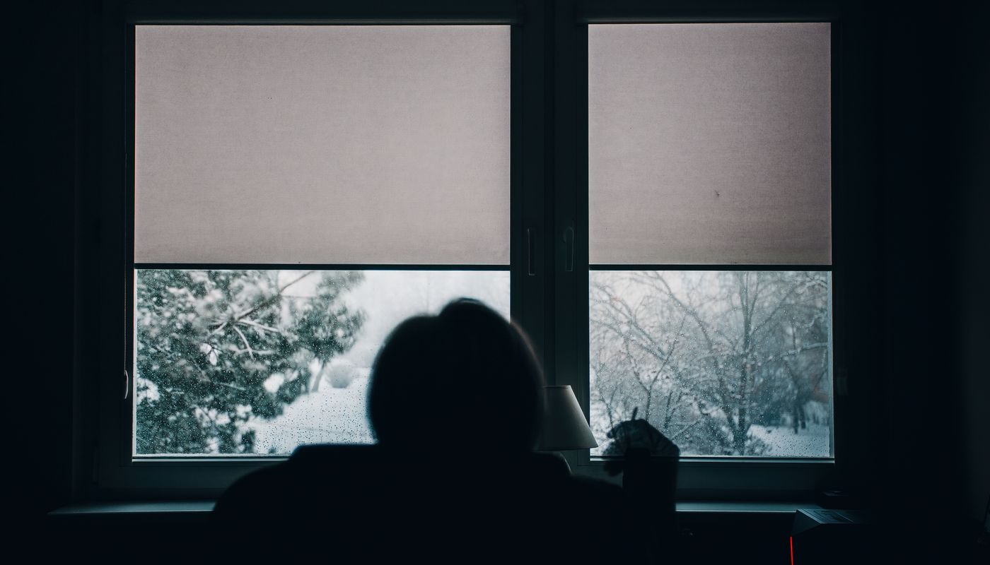 A person sits in silhouette in a dark room facing two windows with half-drawn blinds, looking out at a snow-covered garden