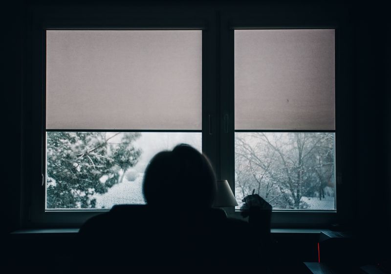 A person sits in silhouette in a dark room facing two windows with half-drawn blinds, looking out at a snow-covered garden