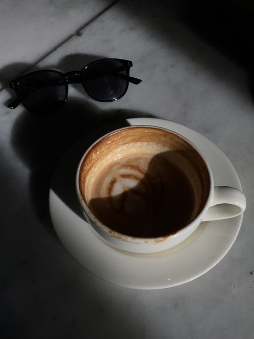 Free A sunlit coffee cup with froth art beside sunglasses on a marble surface. Stock Photo