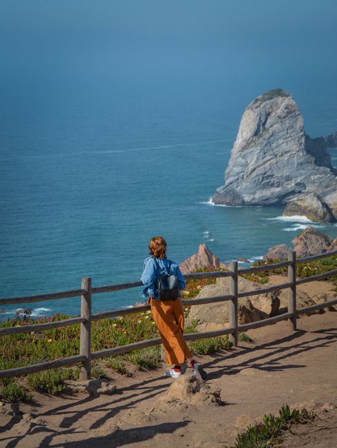 A woman gazes at the vast blue ocean from a cliffside viewpoint on a sunny day.