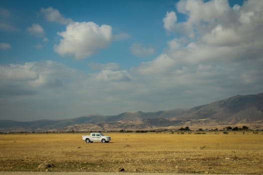 A white pickup truck in a vast grassy field under a cloudy sky, set against Salalah's scenic mountain backdrop.